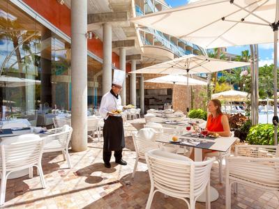 Außenrestaurant mit weißen Möbeln, Sonnenschirmen und Kellner in schwarzer Schürze.