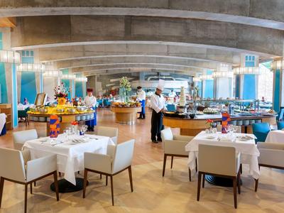 Moderner, heller Speisesaal mit weißen Tischen, bequemen Stühlen und Buffetstationen unter Betonbalken.