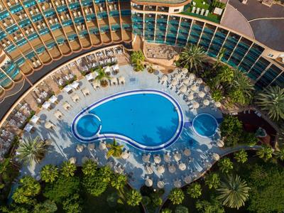 Blick von oben auf einen großen Hotelpool in tropischer Umgebung mit Liegestühlen und Palmen.