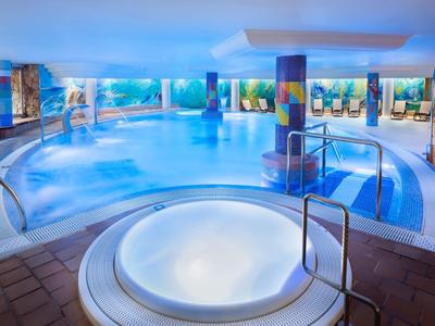 Ein großes Hallenbad mit klarem Wasser, blauem Licht und einem runden Whirlpool im Vordergrund.