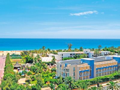 Hotelová budova u moře s zahradami a blízkou pláží pod jasnou modrou oblohou.