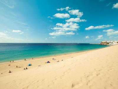 Široká písčitá pláž s málo lidmi a jasně modrou oblohou nad mořem.