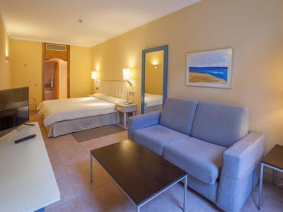 Světelný hotelový pokoj s manželskou postelí, pohovkou, konferenčním stolkem a televizí.