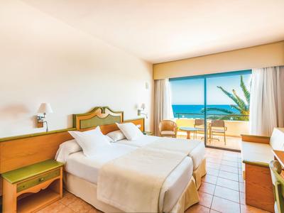 Lichte hotelkamer met tweepersoonsbed, balkon en zeezicht op een zonnige dag.