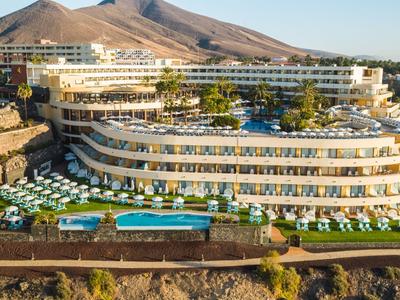 Modern hotel met meerdere zwembaden en ligbedden voor een bergachtige achtergrond.