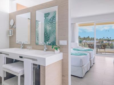 Modern hotelkamer met tweepersoonsbed, badkamer en balkon met uitzicht op palmbomen en zee.