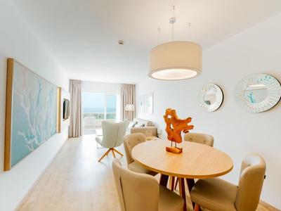 Bright dining room with round wooden table, beige chairs, and seating area with sea view.