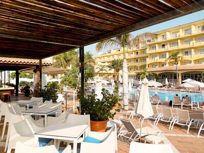 View from covered terrace to hotel pool and multi-story hotel building with palm trees.