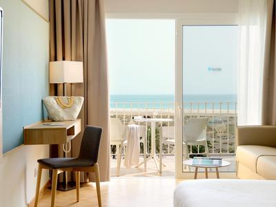 Bright hotel room with desk, chair, sofa and balcony view overlooking the sea.