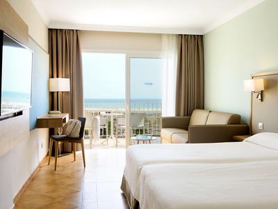 Modern hotel room with two single beds, sofa, desk, and sea view through large windows.