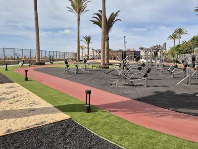 Outdoor-Fitnessbereich mit Cardio-Geräten und Laufbahn in einer sonnigen Hotelanlage nahe Palmen und Meer