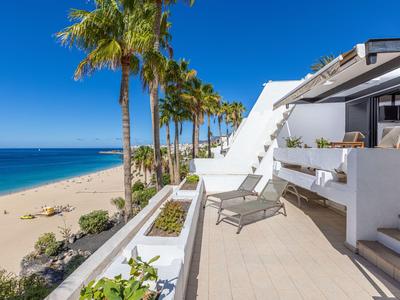Terrasse avec chaises longues donnant sur des palmiers et une plage de sable sous un ciel bleu clair