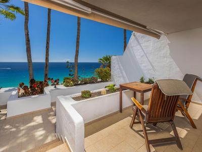 Balcon avec chaise en bois donnant sur la mer bleue et des palmiers sous un ciel clair.