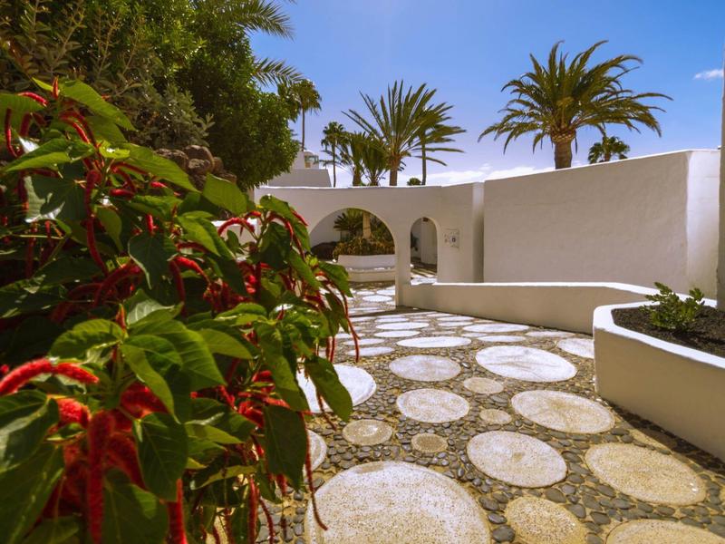 Slunné nádvoří s bílými zdmi, palmami a kulatými kamennými deskami na zemi.