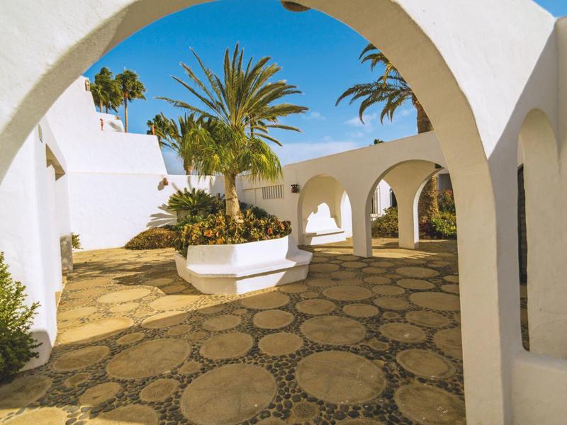 Nádvoří s bílými oblouky, palmami a kamennou podlahou pod modrou oblohou.