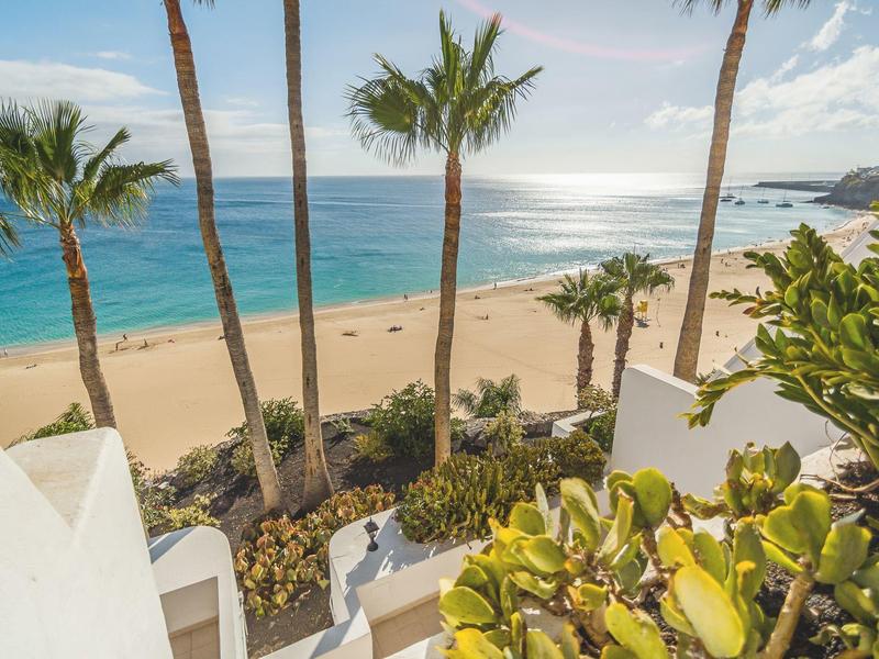 Pohled na písečnou pláž s palmami a bílými budovami u moře.