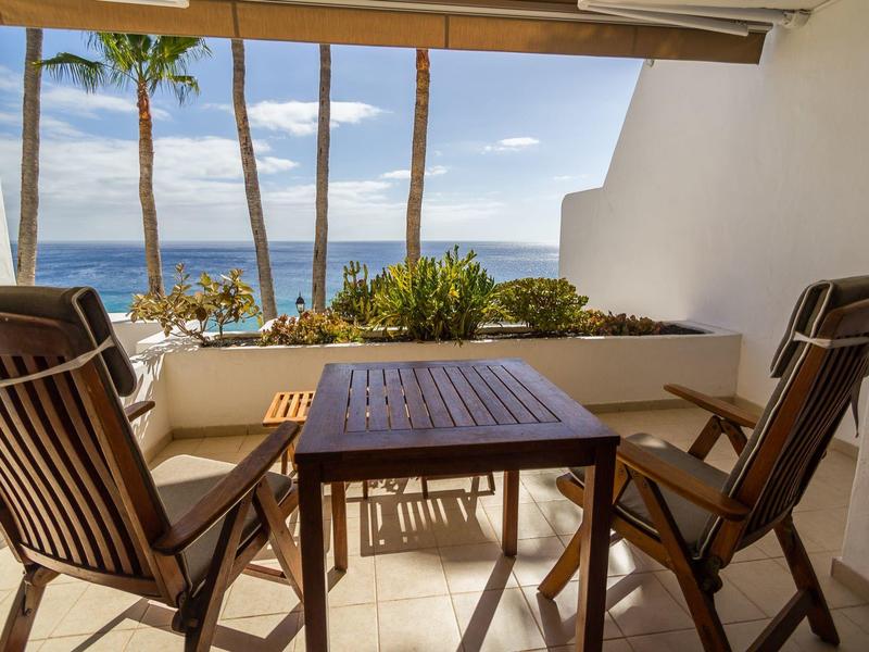 Balkon s dřevěným stolem a židlemi, výhled na palmy a moře pod modrou oblohou.