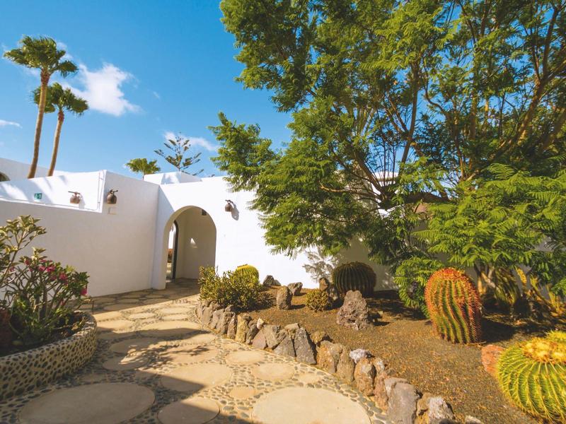 Hotelová zahrada s tropickými rostlinami, kaktusy a bílou budovou pod modrou oblohou.