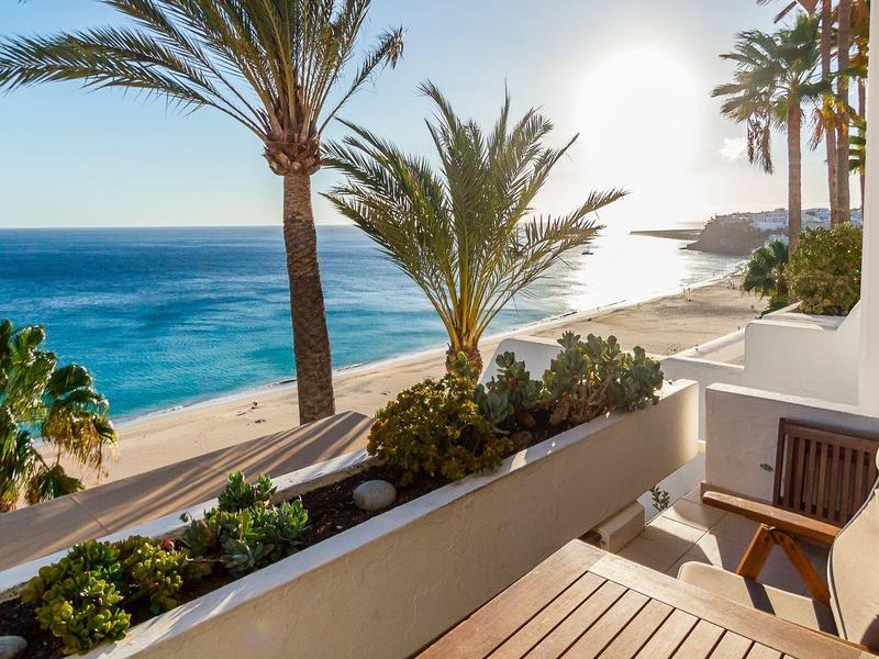 Balkon s židlí a stolem s výhledem na pláž, palmy a moře za slunečného počasí.