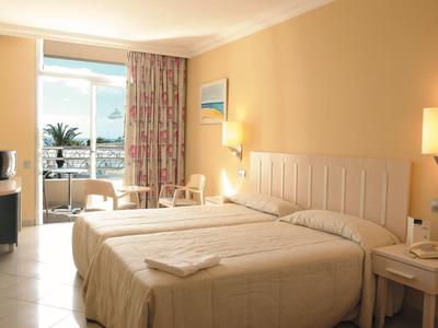 Bright hotel room with two beds, balcony, and view of palm trees and sea.