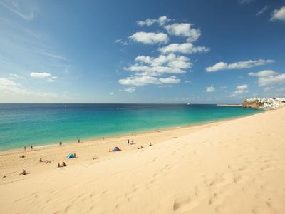 Wide sandy beach with blue sky and scattered clouds, some people near the sea.