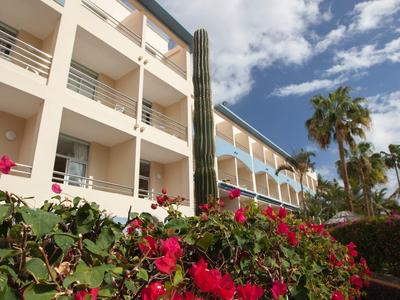 Hotel building with balconies beside red flowers and palm trees under blue sky.