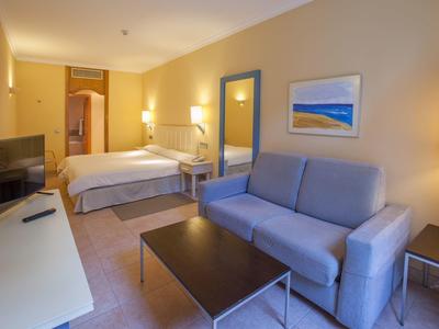 Cozy hotel room area with bed, sofa, table, and TV in warm lighting.