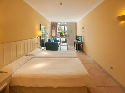 Bright hotel room with two beds, seating area, and balcony access overlooking green plants.