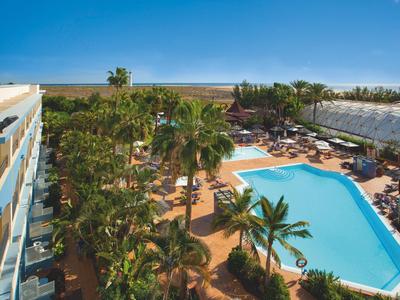 Piscina dell'hotel con palme e ombrelloni, vista sulla spiaggia sotto un cielo soleggiato.