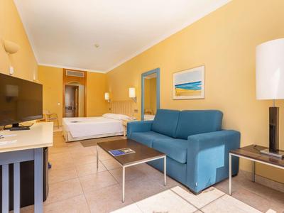 Helles Hotelzimmer mit blauem Sofa, Doppelbett und großem Fernseher.