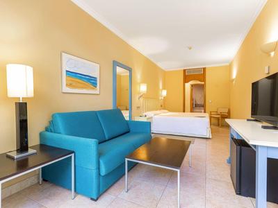 Helles Hotelzimmer mit blauem Sofa, Bett, TV und modernen Möbeln in warmen Farben.