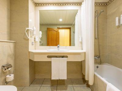 Helles Hotelbadezimmer mit Waschbecken, großem Spiegel, Badewanne und Toilettenpapierhalter.