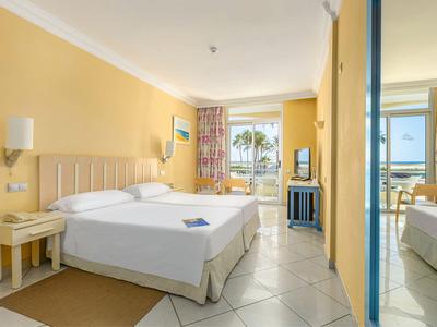 Helles Hotelzimmer mit großem Bett, gelben Wänden und Blick auf Balkon und Meer.