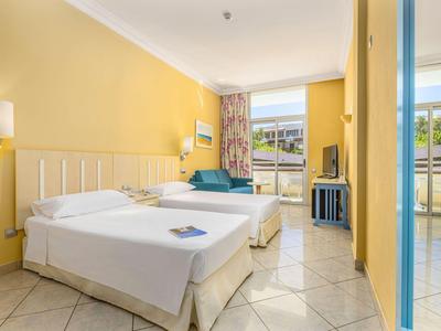 Hellgelbes Hotelzimmer mit Doppelbett, Sofa, Fliesenboden und Balkon mit Blick ins Grüne.