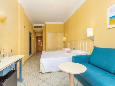 Hotelzimmer mit zwei Einzelbetten, blauem Sofa, rundem Tisch und gelben Wänden.