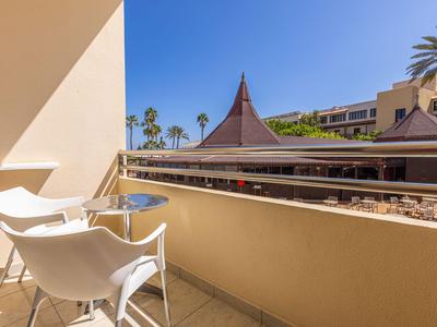 Balkon mit zwei weißen Stühlen und kleinem runden Tisch mit Blick auf Hotelgebäude und Palmen.