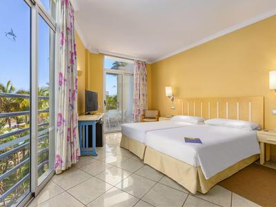 Helles Hotelzimmer mit Doppelbett, großem Fenster und Balkon mit Blick auf Palmen.