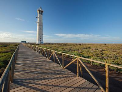 A long wooden boardwalk leads to a white lighthouse in an open landscape under a blue sky.