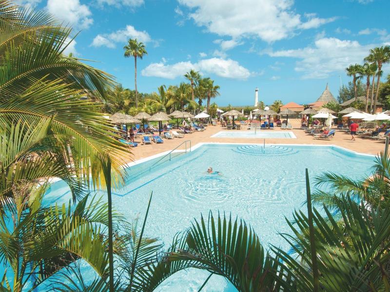 Large pool with palm trees and sun loungers under a blue sky at a hotel resort.
