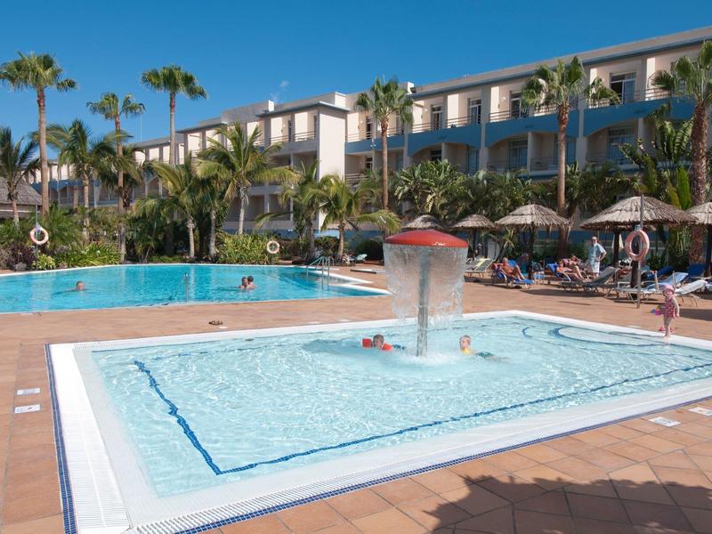Hotel pool area with a children's pool and sun umbrellas under palm trees.