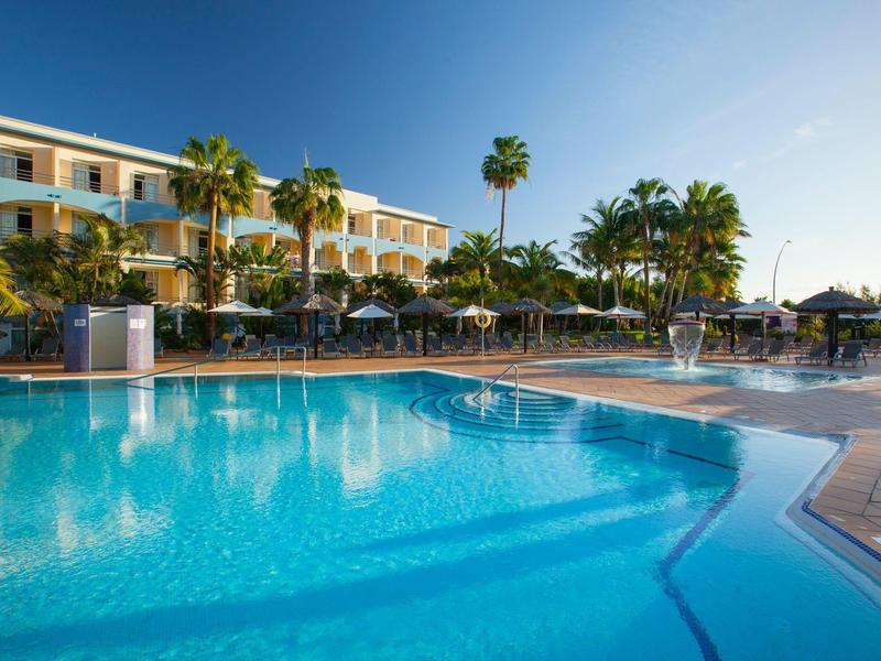 Hotel with large outdoor pool surrounded by palm trees and sun loungers under clear sky.