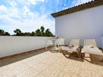 Terrasse mit hellen Fliesen, zwei Liegestühlen und kleinem weißen Tisch unter blauem Himmel.