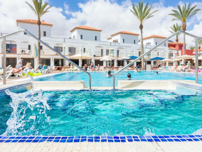 Großer blauer Pool mit Sprudelliegen, Sonnenschirmen und weißen Gebäuden im Hintergrund.