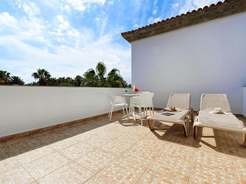 Terrasse mit hellen Fliesen, zwei Liegestühlen und kleinem weißen Tisch unter blauem Himmel.