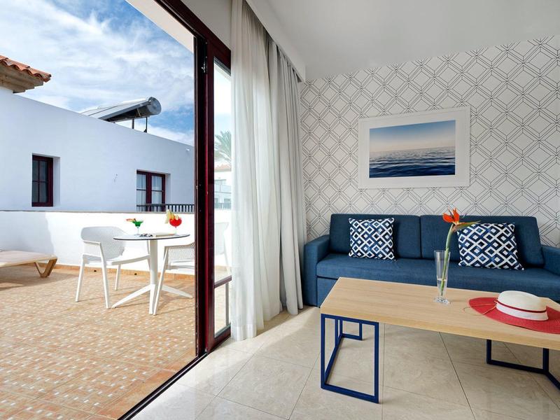 Heller Wohnraum mit blauer Couch, Holz-Couchtisch, weißem Vorhang und Zugang zur Terrasse mit Möbeln.