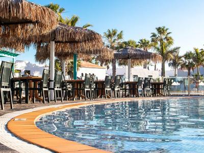 Zwembadgedeelte met stro parasols en stoelen naast palmbomen onder een heldere lucht.
