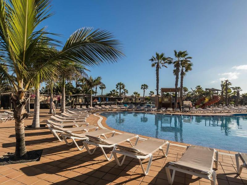 Large hotel pool with sun loungers and palm trees under a clear blue sky.