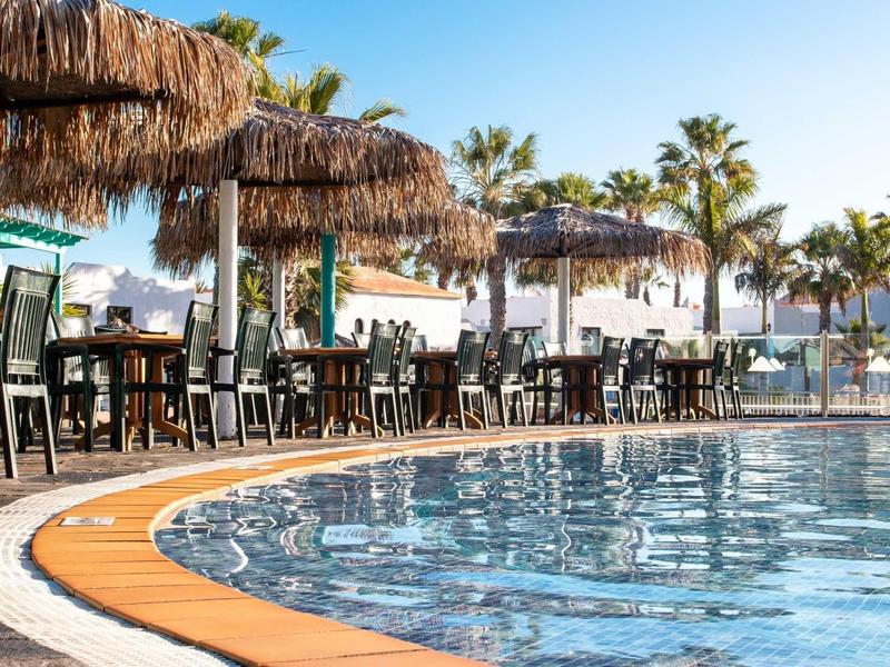 Pool area with loungers, tables, and thatched umbrellas by the water