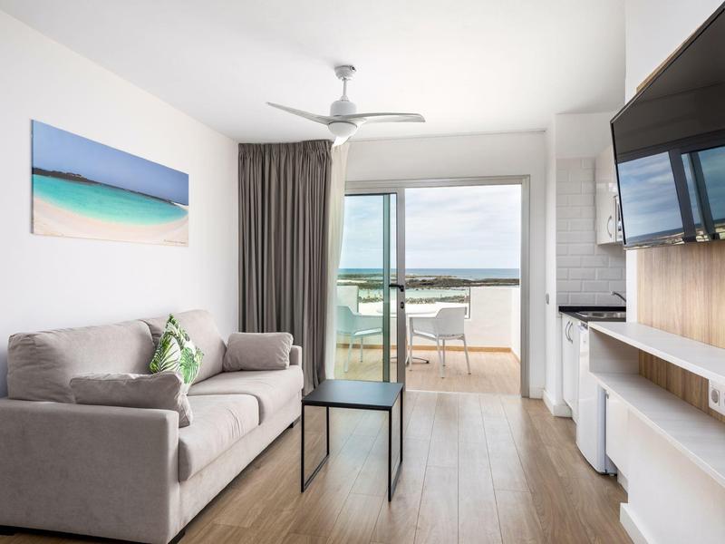 Moderner Wohnraum mit grauem Sofa, TV, hellem Holzboden und Balkon mit Meerblick.