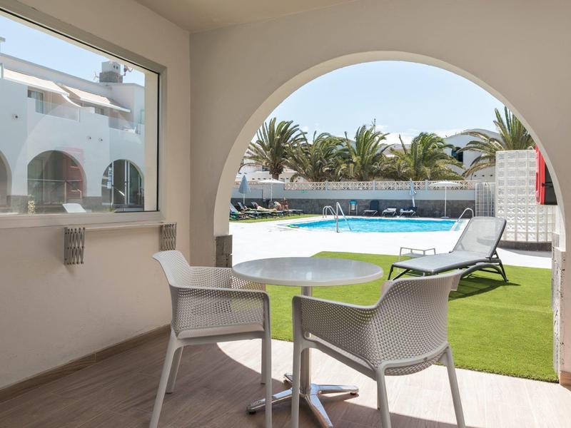 Blick durch Rundbogen auf Terrasse mit Tisch und Stühlen, Pool und Palmen im Hintergrund.